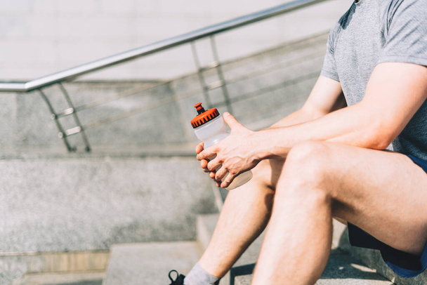 Nahaufnahme eines müden jungen Mannes, der auf einer Treppe sitzt und sich nach dem Sport entspannt. Halten einer Wassermixer-Flasche während des Trainings in der sommerlichen City Street, bewölkter Himmel - Foto, Bild