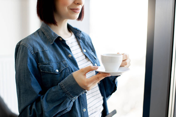 Close up of pleasant caucasian woman in casual wear holding cup of hot beverage while standing at office. Panoramic window on blurred background. Short coffee break before work. - Φωτογραφία, εικόνα