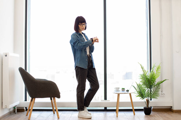 Full length portrait of caucasian dark haired woman standing near panoramic window and looking on smartphone wearing on wrist. Office worker in casual attire using app on portable gadget. - Фото, изображение