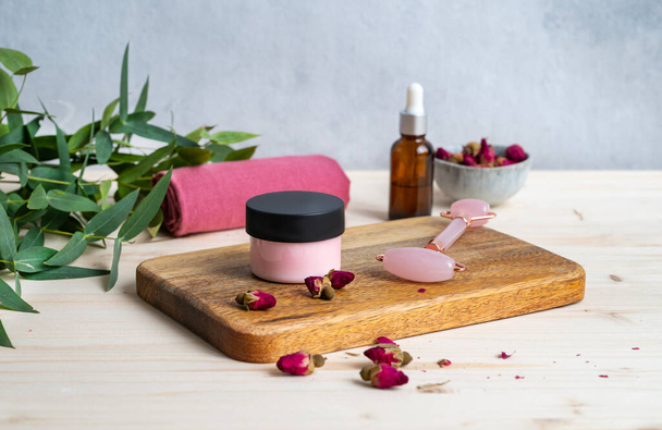 The concept of face and body care cosmetics with rose and eucalyptus. Face cream and massager on a wooden table with dried rose flowers. - Photo, Image