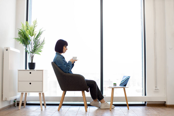 Dark haired woman in casual wear and glasses sitting in office armchair near panoramic window and enjoying hot aroma coffee. Small coffee table with portable laptop standing in front of female worker. - Foto, Bild