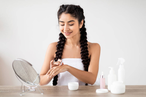 Skincare Cosmetics. Young beautiful indian woman applying hand cream at home, smiling eastern female moisturizing skin after bath, sitting wrapped in towel at dressing table in bedroom - Фото, зображення