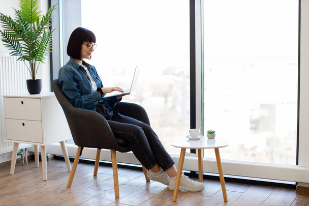 Attractive dark haired woman sitting in armchair with wireless laptop in hands and looking at amazing view from panoramic window at office. Casually dressed brunette enjoying her creative workplace. - Foto, Imagem