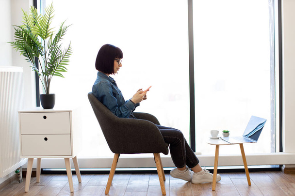 Pleasant dark haired woman typing messages on smartphone while sitting in comfy office chair. Caucasian female employee in eyeglasses chatting with coworkers online using modern gadget. - Foto, afbeelding