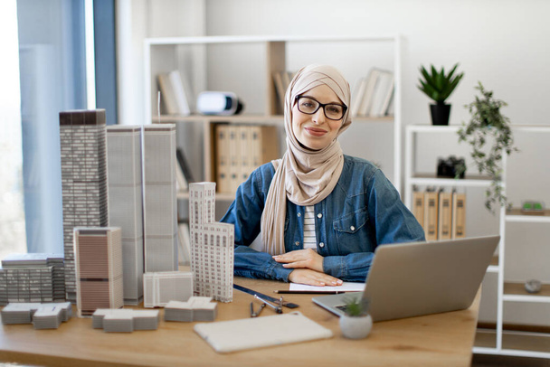Young muslim woman wearing hijab and spectacles sitting at office desk in real estate agency. Efficient realtor using wireless technologies while arranging purchase of residential properties indoors. - Photo, image