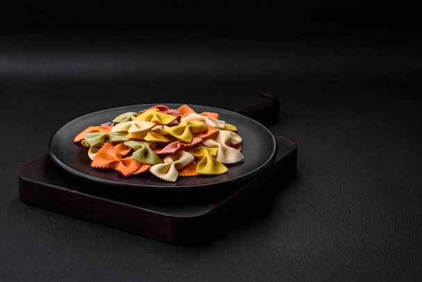 Pasta de farfalle cruda en diferentes colores sobre un fondo de hormigón oscuro. Preparación para cocinar comida italiana - Foto, Imagen