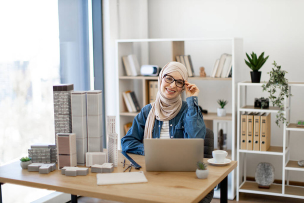 Cheerful muslim lady in hijab and casual clothes touching glasses while smiling behind writing desk in spacious workplace. Successful employee creating architectural design in office interior. - Foto, afbeelding