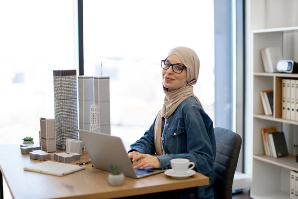 Architecte musulmane en hijab travaillant sur ordinateur portable au bureau avec des modèles de construction dessus. Professionnel de la conception expérimenté menant l'urbanisme en utilisant des technologies avancées. - Photo, image