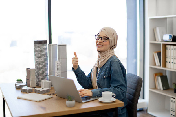 Muslim female architect working on portable computer at office desk with building models on it showing thumb up. Experienced design professional conducting city planning using advanced technologies. - Foto, Imagen