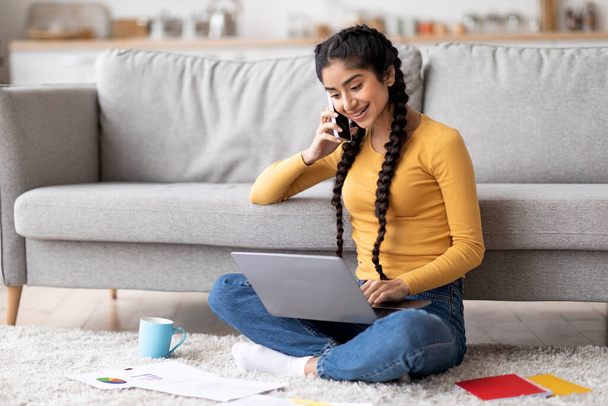 Ινδή ελεύθερος επαγγελματίας γυναίκα που χρησιμοποιεί το lap-top και μιλώντας στο κινητό τηλέφωνο στο σπίτι, χαμογελώντας ανατολικές γυναίκες που εργάζονται εξ αποστάσεως με τον υπολογιστή και τα έγγραφα, ενώ κάθεται στο πάτωμα στο σαλόνι, αντιγράψτε το διάστημα - Φωτογραφία, εικόνα