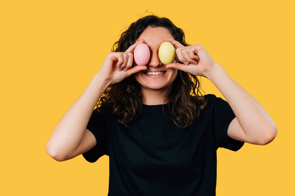 Vidám fiatal mosolygós nő eltakarja a szemét színes húsvéti tojások sárga háttér. - Fotó, kép