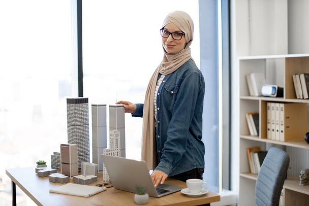 Happy muslim lady wearing beige hijab using drafting compass while working with architectural models in office. Efficient design professional comparing measurements with digital data on computer. - Foto, Imagen