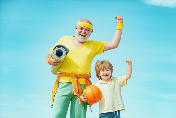 Senior man and child exercising on blue sky. Sports for kids. Sportsman grandfather and healthy kid with basketball ball on blue sky background - Foto, Imagem