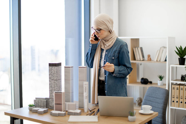 Smiling arabian woman in beige hijab holding hand with pencil on building model while talking on phone in creative workplace. Talented professional having business conversation in architecture office. - Zdjęcie, obraz