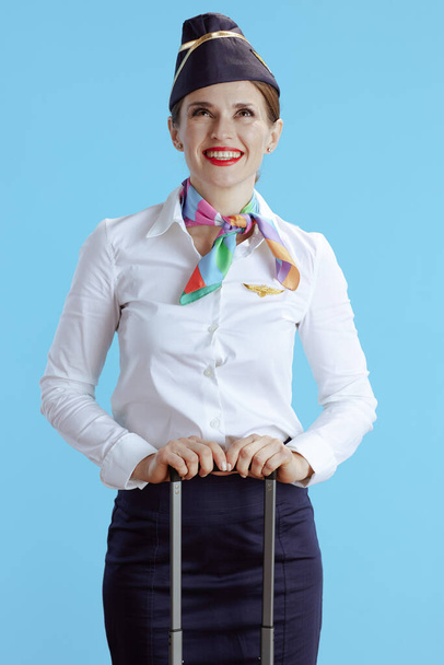 smiling elegant female flight attendant isolated on blue background in uniform with travel bag looking up at copy space. - Photo, image