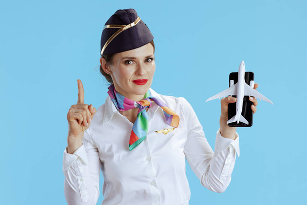 smiling modern female stewardess against blue background in uniform with a little airplane and smartphone with raised finger drawing attention. - Fotó, kép