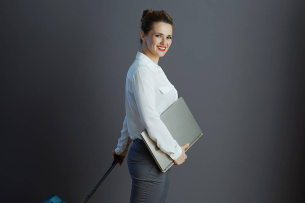 smiling 40 years old business woman in white blouse with laptop and trolley bag against gray background. - Photo, Image