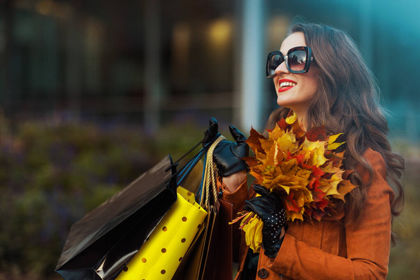 Hola otoño. feliz mujer moderna de 40 años en gabardina naranja con bolsas de compras y hojas amarillas de otoño cerca de la tienda en la ciudad. - Foto, imagen