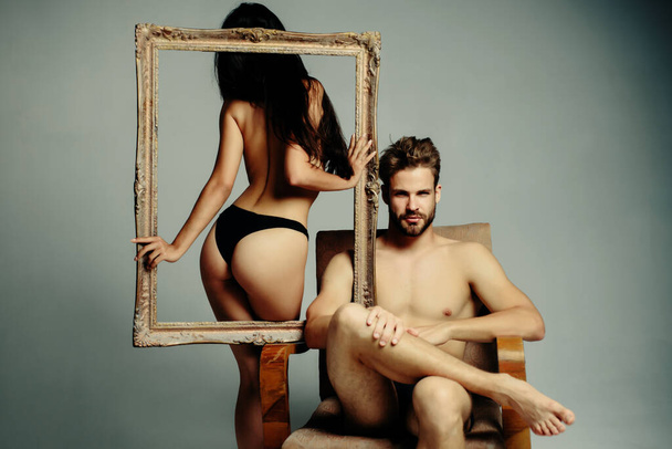 Beautiful sexy couple. Young woman holding an empty frame. Brunette girl in a black panties with guy with naked torso. Confident brutal man. Isolated gray background - Φωτογραφία, εικόνα