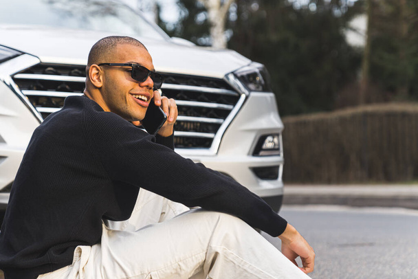 Αφροαμερικάνος με γυαλιά ηλίου μιλάει στο τηλέφωνο, κάθεται σε ένα πεζοδρόμιο μπροστά από ένα αυτοκίνητο. Υψηλής ποιότητας φωτογραφία - Φωτογραφία, εικόνα