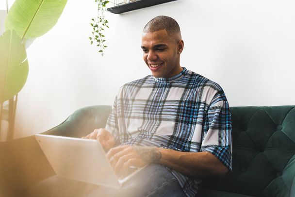 Усміхнений афроамериканець друкує на ноутбуці, сидячи вдома на дивані. Фотографія високої якості - Фото, зображення