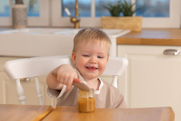 家庭で幸せな家族。子供が台所で食事をしている。乱雑な面白い顔をした小さな男の子は健康的な食べ物を食べる。子供はスプーンを持って一人で食べる。自給自足 - 写真・画像