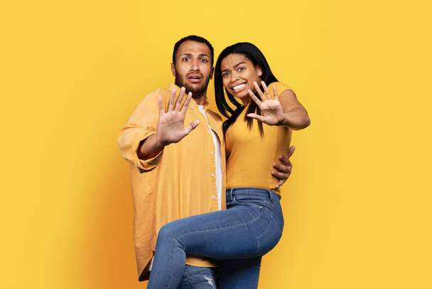Frightened millennial african american male and wife making stop hand gesture afraid isolated on yellow studio background, copy space. Fear, horror people emotions, disgust sign - Photo, Image