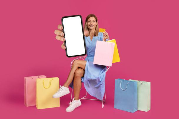 Šťastný středního věku evropské blondýny žena s mnoha balíčky s nákupy ukazuje smartphone s prázdnou obrazovkou izolované na fialové pozadí studia. Prodej, aplikace pro online nakupování - Fotografie, Obrázek
