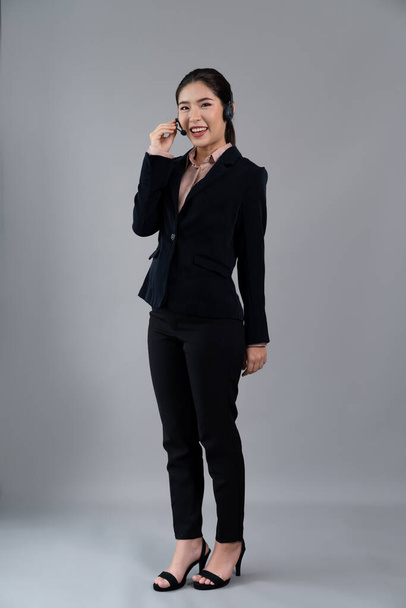 Attractive full body asian female call center operator with happy face advertises job opportunity on empty space, wearing formal suit and headset on customizable isolated background. Enthusiastic - Foto, afbeelding