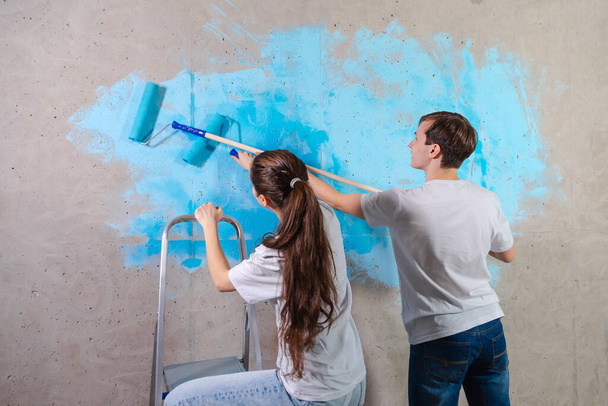 Pár v novém domově při opravách malování stěn společně. Šťastná rodina drží barvu váleček malování stěnu s modrou barvou barvy v novém domě. Domácí renovace DIY obnovit domácí koncept - Fotografie, Obrázek