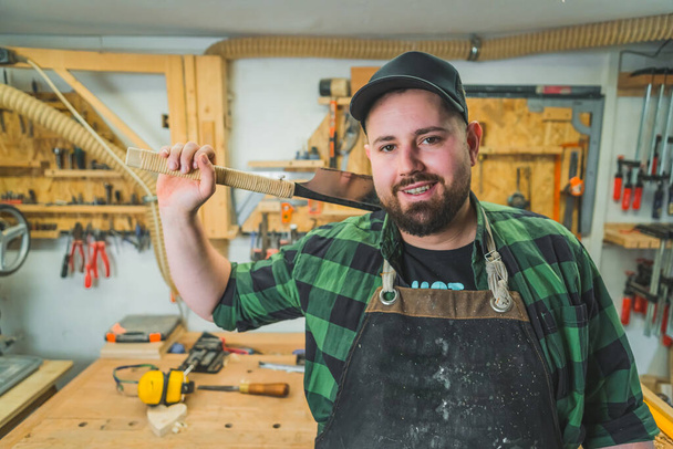 Koncept dřevoobráběcích nástrojů. Pozitivní muž, dospělý tesař, drží jeden ze svých nástrojů a s úsměvem se dívá do kamery. Interiér dílny. Kvalitní fotografie - Fotografie, Obrázek