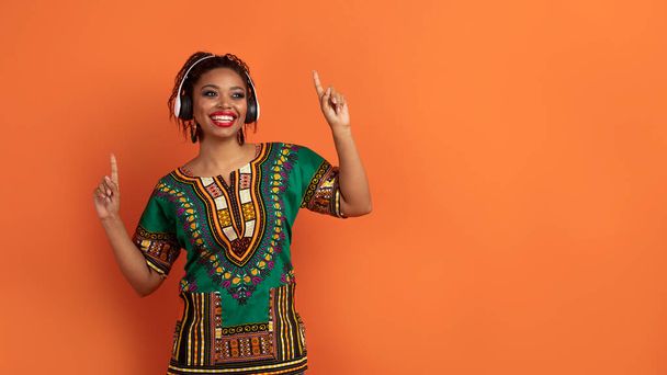 Joyeux joyeux millénaire dame noire en costume africain traditionnel lumineux en utilisant des écouteurs sans fil, écouter de la musique et de la danse, en regardant l'espace de copie pour la publicité sur fond de studio orange - Photo, image