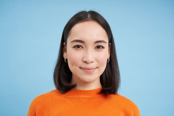 Close up of candid young asian woman, looking at camera with genuine, natural emotions, standing over blue background. - Фото, зображення