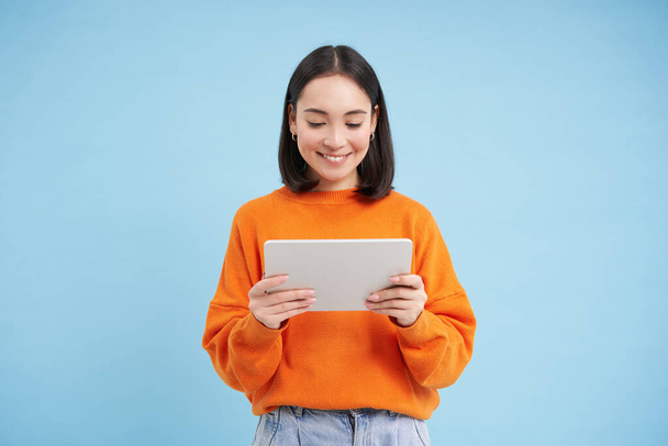 Χαμογελώντας όμορφη Ασιάτισσα που διαβάζει σε ψηφιακό tablet, βλέποντας βίντεο σε gadget ή παίζοντας βιντεοπαιχνίδια, στέκεται πάνω από το μπλε φόντο. - Φωτογραφία, εικόνα