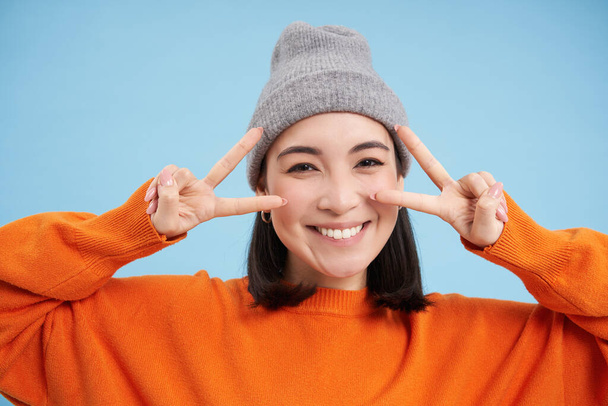 Close up portrait of cute korean girl in beanie, shows peace, vsign gesture, positive vibe, smiling and laughing, posing over blue background. - Valokuva, kuva