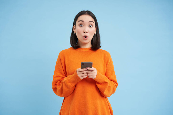 Удивительная корейская девушка держит смартфон, говорит вау, выглядит впечатлен мобильным приложением предложение, продвижение сайта, стоит на синем фоне. - Фото, изображение