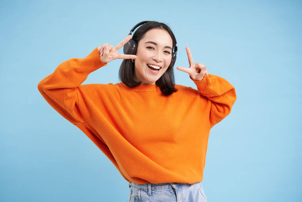 Красива корейська жінка в навушниках, танцює і слухає музику в навушниках, стоячи на синьому фоні
. - Фото, зображення