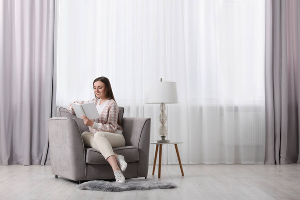 Woman reading book in armchair near window with stylish curtains at home. Space for text - Foto, Imagem