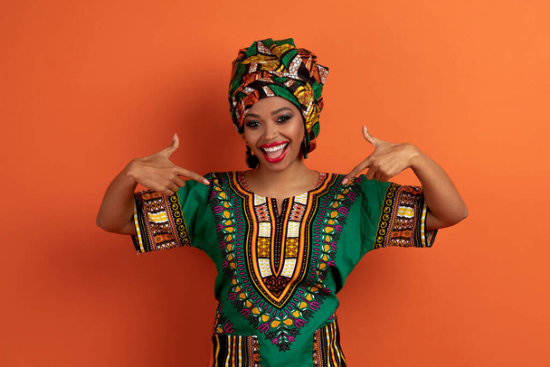 Etnisyys, kulttuuri, Afrikka käsite. Iloinen onnellinen positiivinen kaunis nuori musta nainen yllään perinteinen afrikkalainen vaatteet ja kirkas meikki osoittaa itseään ja hymyilevä, oranssi studio tausta - Valokuva, kuva