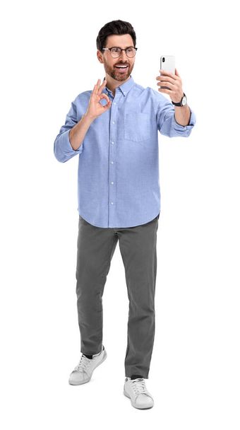 Smiling man taking selfie with smartphone and showing OK gesture on white background - Foto, Imagen