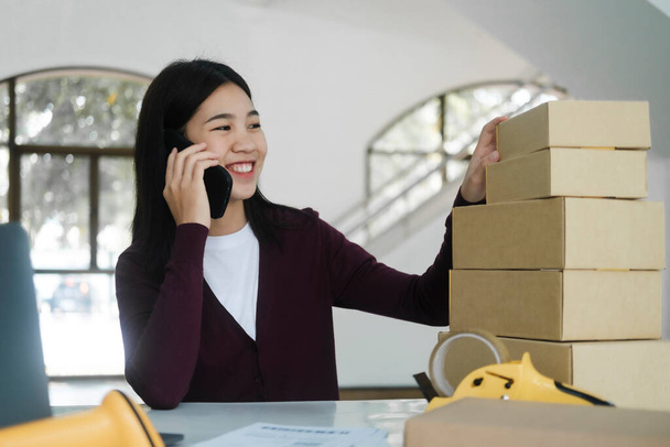 Young asian female online business owner or entrepreneur with smiley face sitting at desk with boxes, laptop, scanner, and taper talking to customer for order using smartphone. Online business, e-commerce concept. - Photo, Image