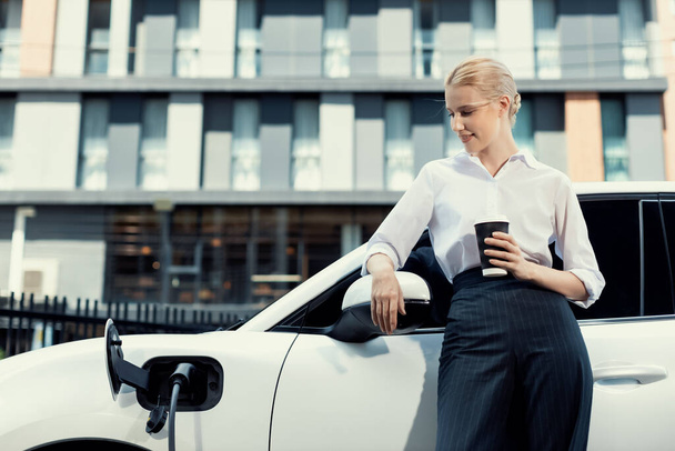 Businesswoman drinking coffee, leaning on electric vehicle recharging at public charging station with residential apartment condos building in background as progressive lifestyle by eco-friendly car. - Photo, image