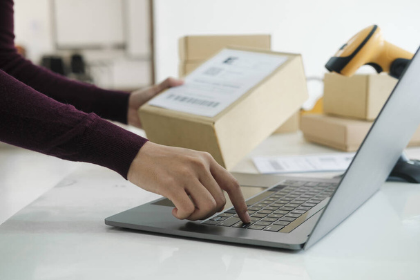 Close up of female online business owner, entrepreneur holding parcel while standing and checking customer's information with laptop before shipment or delivery to customer. Online business, e-commerce, logistics concept. - Photo, Image