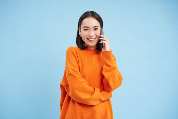 Korean girl in orange sweater talks on mobile phone, speaks on cellphone with happy smiling face, orders delivery, consults with someone on telephone. - Fotografie, Obrázek
