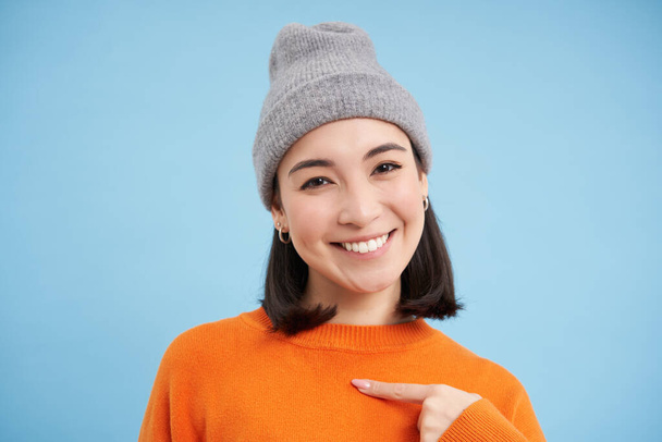 Közelkép portré koreai lány téli kalap, rámutat magára, áll narancssárga pulóver felett kék háttér. - Fotó, kép