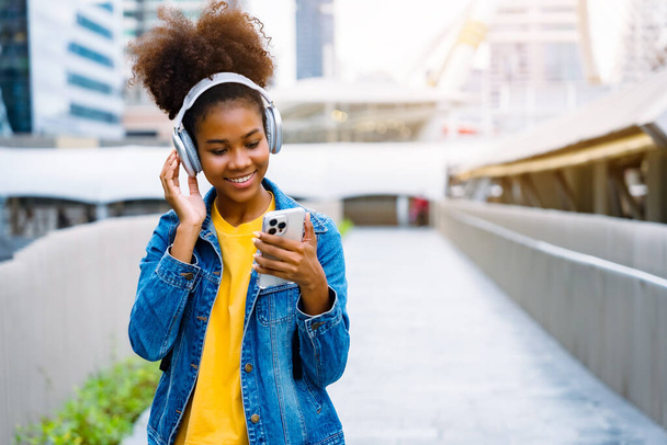 Menina estudante negro alegre usando fone de ouvido e usando smartphone móvel, Caminhando no prédio da faculdade ao ar livre, Feliz e sorridente adolescente mulher. Educação moderna - Foto, Imagem