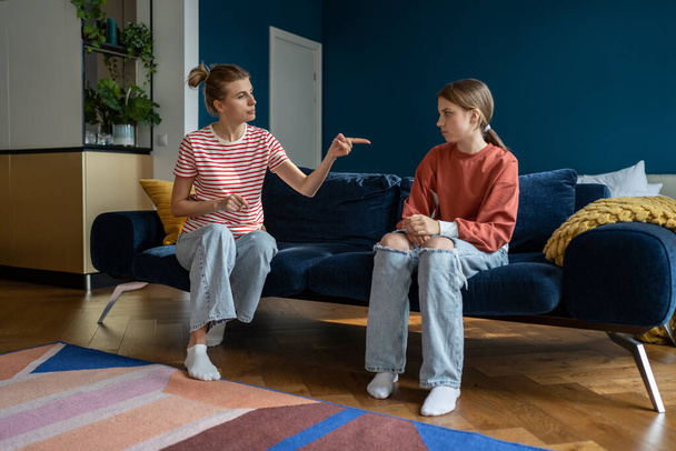 Parent-teen conflict. Strict mother scolding lecturing upset teenage girl daughter at home, pointing finger. Mom disciplining teenager while sitting together on sofa. Parenting adolescent - Photo, image