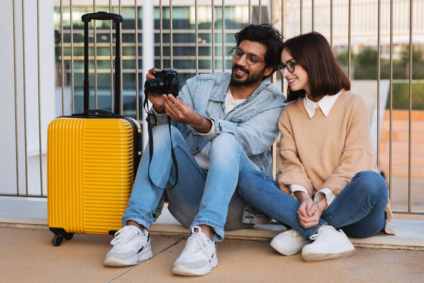 Smiling millennial indian guy and caucasian female tourists sitting with yellow suitcase and looking at photo on camera in city outdoor. Travel blog, vacation and trip together - Photo, Image