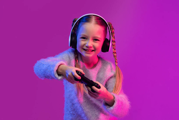 Happy sweet blondy preteen girl playing video games on colorful background in neon light, using wireless headphones and holding joystick. Game addiction for children concept - Foto, Bild
