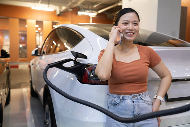 A young woman and her electric car at electric charging station in city center . EV urban lifestyle concept - Photo, Image